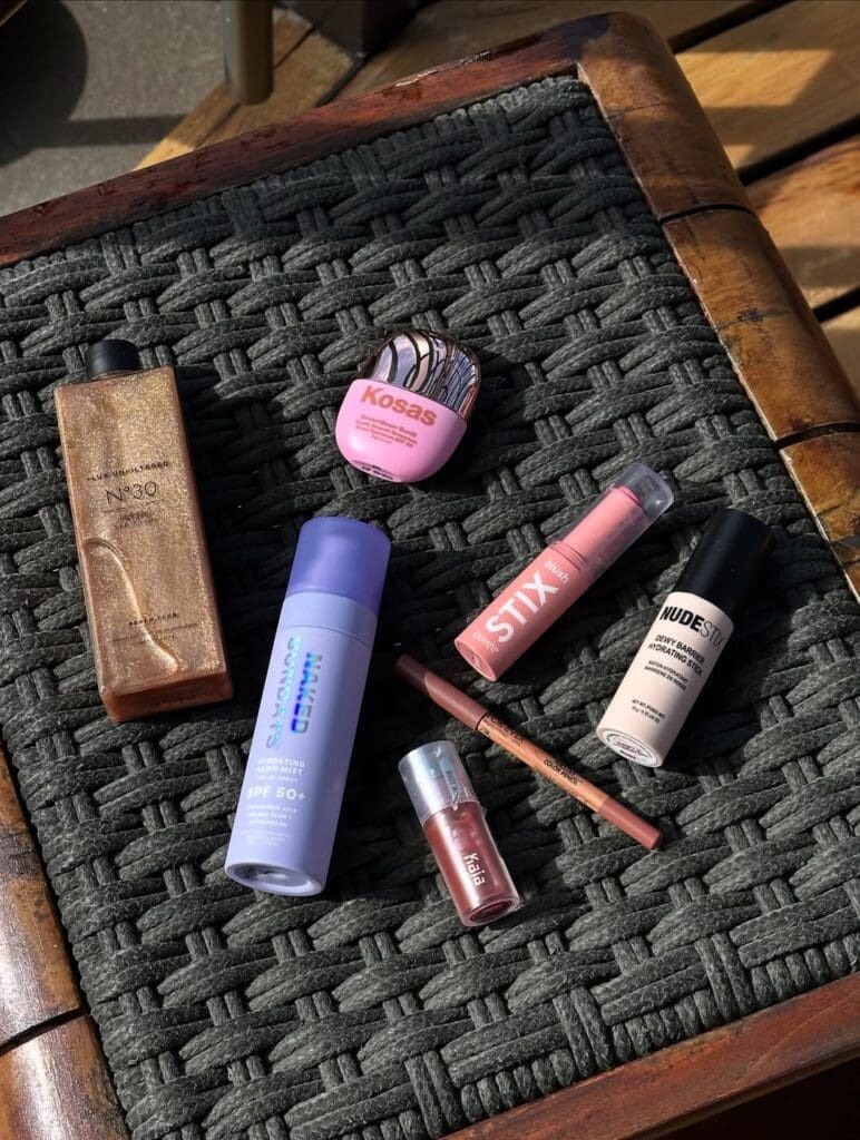 Flat lay of travel beauty essentials including sunscreen, lip products, and shimmer oil on a woven tray.