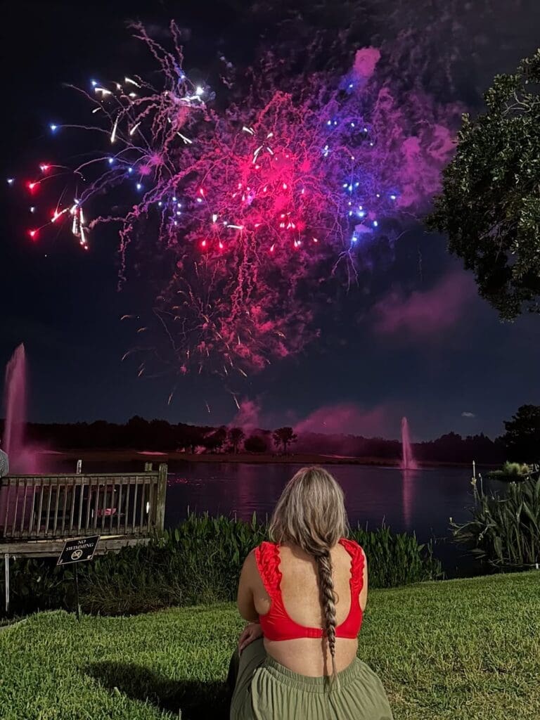 Fireworks show over the lake at JW Marriott Orlando with a woman in a red top watching from the grass.